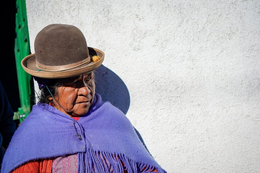 Portrait of a woman in a bowler hat and purple shawl against a white wall